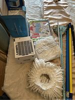 Two Arctic Air coolers, one in original box with USB cable, the other unit missing power cord, adjustable clothes folding board with instructions, and microfiber ring mop head on bed.
