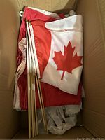 Box containing one large Canadian flag and multiple small hand-held Canadian flags on wooden sticks, all folded and laid inside.