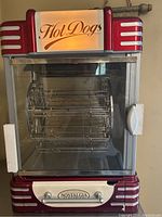 Front view of the Nostalgia Electrics hot dog cooker showing the illuminated 'Hot Dogs' sign, glass enclosure, metal rotating racks inside and sliding storage drawer.