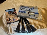 4 black tapered bed risers, Duramax rug gripper new in box, and Comfort Zone window insulation kit in its packaging placed on a fabric surface.