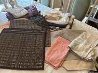 Group of assorted folded linens including tablecloths, napkins, placemats, and runner in various neutral and soft colors, displayed on a table.
