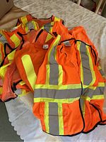 Five fluorescent orange safety vests and one fluorescent orange t-shirt laid out on a surface, showing different styles and reflective striping.