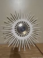 Clear frontal photo of a vintage starburst wall clock showing white wooden spokes radiating from a metal face with Roman numerals.