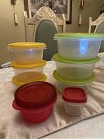 Image of all 8 plastic food storage containers with lids on a table, showing three green lid containers stacked, three yellow lid containers stacked, one medium red lid container, and one small red lid rounded square container.