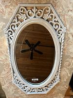 Front facing view of white framed oval mirror with ornate detailing showing the ceiling fan reflection and small crack in glass.