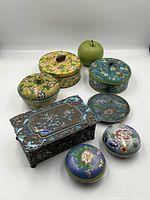 Overview of eight cloisonné pieces arranged on white background with green apple for scale
