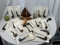 Wide shot showing twenty blister-packaged pipes arranged around a green apple reference and the wooden sail-motif stand.