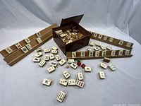 View of bone and bamboo Mah Jong tiles arranged on wooden tile racks with tiles scattered in front and wooden box behind.