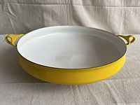 Front and top angled view of yellow Dansk paella pan showing round shape, white enamel interior, yellow enamel exterior, and integrated loop handles.