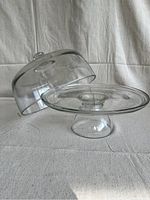 Glass cake stand base and dome cover placed side by side on a neutral fabric surface.