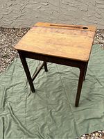 Full view of the wooden school desk showing the top surface and legs placed on a green mat outdoors on gravel.