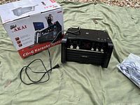 Akai karaoke machine with box, power cord, and microphone laid out on green tarp.
