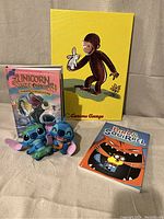 Curious George artwork, Unicorn Rescue Society book, Bird and Squirrel book, and two Stitch plush toys displayed together