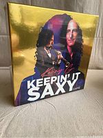 Front view of the Kenny G Keepin' It Saxy game box showing the gold glossy finish and images of Kenny G with saxophone.