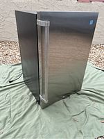 Front side view of the Vissani outdoor mini-fridge showing stainless steel door and handle on green cloth background outdoors.