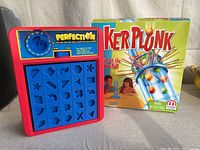 Photo showing Perfection game board with yellow pieces and Kerplunk boxed game standing next to it.