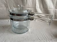Side view of vintage Pyrex double boiler showing clear glass pots with metal banding, clear glass handles and lid.
