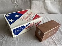 Photo showing the white All American Trivial Pursuit card set box and smaller brown Welcome to America card set box side by side.