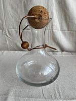 Front view of the clear glass decanter showing round base, narrow neck, cork stopper attached with leather cord and wooden bead.
