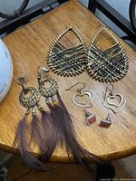 Photo of three pairs of earrings: gold and black striped teardrop, gold filigree round with feathers, silver wire heart with red stones.