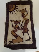 Photo showing rectangular African cloth with depictions of two tribal figures in earth tones on beige fabric with dark border.