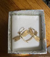 Photo showing a pair of gold tone dragonfly earrings placed inside a white cushioned display within a holographic silver decorative box. The earrings depict dragonflies with detailed wings and segmented tails.