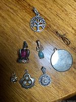 Eight assorted pendants spread on wooden surface showing various shapes and materials: tree of life cutout, pink crown, turquoise stone, silver round locket, pearls, floral cluster, and circular pearl pendant