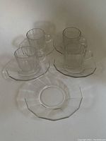 Four clear glass espresso cups each paired with a matching hexagonal-shaped espresso plate, arranged on a flat surface.