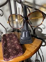 Front view showing vintage tortoiseshell-style large round frame eyeglasses on black display stand alongside brown soft fabric eyeglass case with small floral pattern.