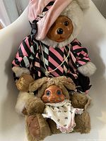 Two vintage Raikes Bears displayed on a white chair, showing the larger bear dressed in pink and black with fur hood and pearl necklace, and the smaller bear wearing bunny ears hat and bib.