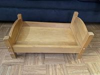 Angled side view of the small wooden doll bed showing the footboard and side panels on wood flooring.