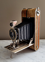 Side front angle showing camera bellows extended, front lens, leather and metal casing, and fold-out stand deployed.