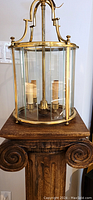 Full view of brass hanging lantern on wooden pedestal showing curved glass panels and candle-style lights inside.
