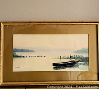 Full view of the framed watercolor painting depicting a calm lake with a boat, stones, reeds, and distant trees.