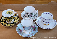 Four assorted vintage fine china cups and saucers on cardboard, floral designs with gold trim and scalloped edges.