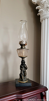 Full view of oil lamp showing glass chimney, etched glass reservoir, and figural base on black platform.