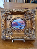 Front view of an ornate giltwood frame with a colorful oval coastal scene print in the center. The frame has floral embossed decorations on all four corners with gold and silver tones.