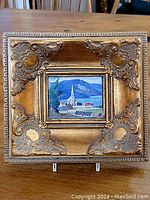 Front view of the small giltwood frame with detailed ornamental corners and a landscape print inside showing a church steeple and mountain scene.