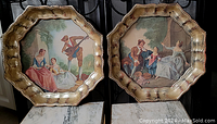 Pair of gold-toned scalloped ornate frames with glass covering showing two romantic pastoral scenes in 18th-century style clothing.