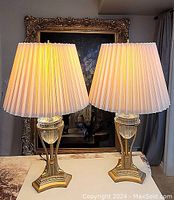 Pair of lamps lit on table, showing ivory pleated shades and gold-tone bases