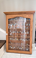 Front closed view of oak display cabinet showing rows of collectible souvenir spoons behind glass door.