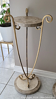 Full view of the tall faux marble and gold metal stand placed on tiled floor near a wall and plant.