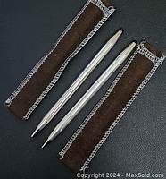 Two sterling silver Cross writing instruments, ballpoint pen and mechanical pencil, laid next to their brown stitched fabric sleeves on a black background.