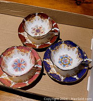Top-down photo showing three tea cups each on their matching saucers inside a cardboard box. Colors visible are deep red, royal blue, and pink with gold and floral designs.