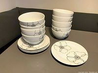 Set of white ceramic bowls and plates with black floral pattern stacked on a table and couch background