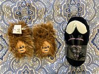 Top view of the two different slippers; brown fuzzy pair with Chewbacca faces, black pair with lightsaber design on soles.