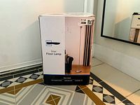Photo of the original white box showing the image and description of the silver floor lamp, labeled as new.