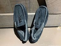 Top view of grey moccasin style slippers showing soft fabric material, stitch detailing, and bow accents on top.