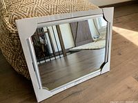 Silver frame rectangular mirror placed on wood floor near a pouf. Shows frame detail and reflection.