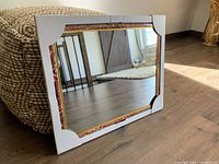 Rectangular mirror with white marble-look frame, gold and brown trim, resting against a woven ottoman on brown wooden floor with sunlight reflection